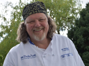 Smiling Bill Ranniger wearing a beanie and Duke's Seafood chef robe
